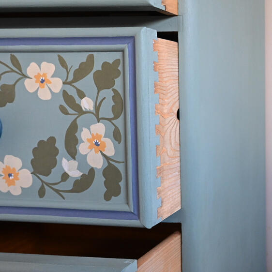 Close-up of a light blue painted drawer with hand-painted floral designs in white, yellow, and green. The drawer has a round blue knob. The drawer is framed by a darker blue trim. There are painted dovetail joints