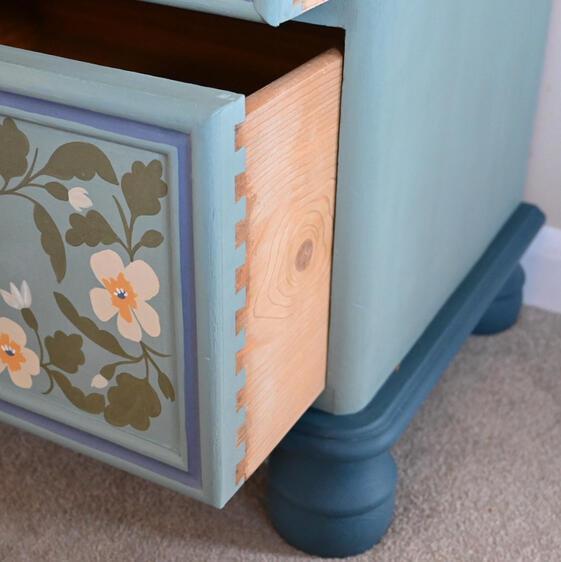 Close-up of a light blue painted drawer with hand-painted floral designs, partially open to reveal the wooden interior with dovetail joints. The drawer has a round blue knob and is framed by darker blue trim. The chest of drawers has a rounded base and fee