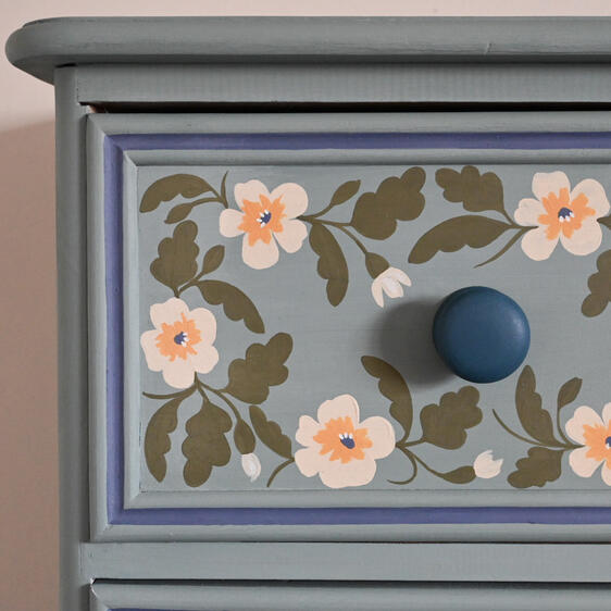 Close-up of a light blue painted drawer with hand-painted floral designs in white, yellow, and green. The drawer has a round blue knob. The drawer is framed by a darker blue trim.