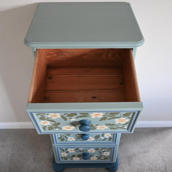 Top-down view of a light blue painted chest of drawers with two drawers showing. The top drawer is open, revealing a light wood interior. Both drawers have hand-painted floral designs in white, yellow, and green, and round blue knobs. The chest has a round