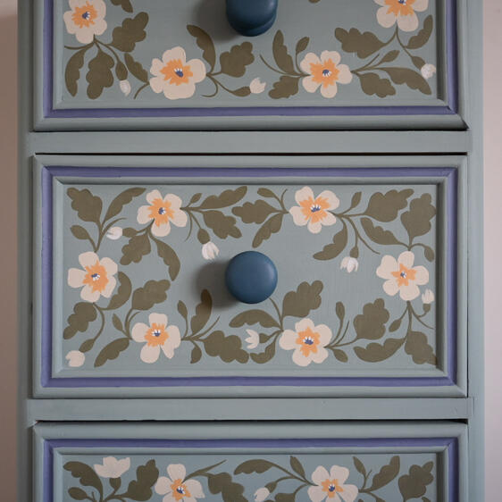 Close-up of three drawers from a chest of drawers, each painted light blue with hand-painted floral designs in white, yellow, and green. Each drawer has a round blue knob.