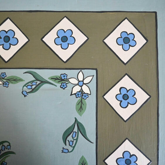 Chalk painted table top Close-up of a hand-painted floral tabletop featuring blue forget-me-not flowers, white blossoms, green foliage, and a decorative geometric border on a vintage duck egg blue painted wooden table.