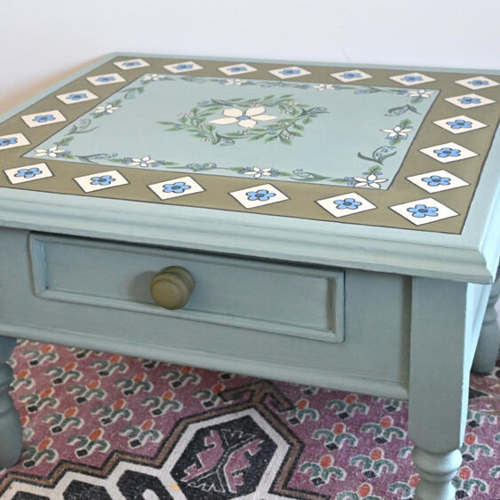 Hand painted floral table Hand-painted vintage blue wooden side table with a single drawer, featuring an elegant floral design and decorative border on the tabletop, refinished in a soft duck egg blue chalk paint.