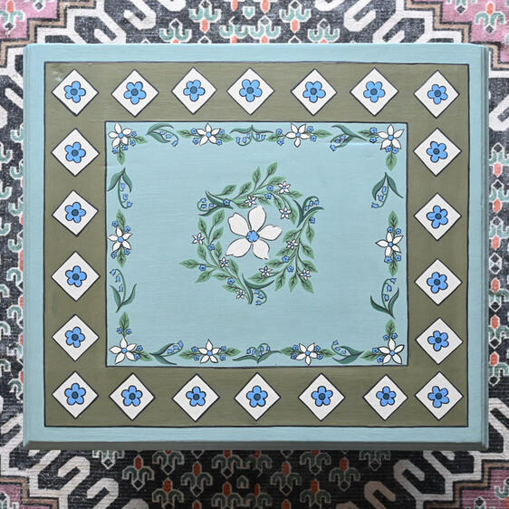 Bird eye view of painted coffee table Overhead view of a hand-painted vintage wooden side table in duck egg blue, featuring a central white floral wreath, delicate blue flowers, and an olive green geometric border with hand-painted blue flower motifs.