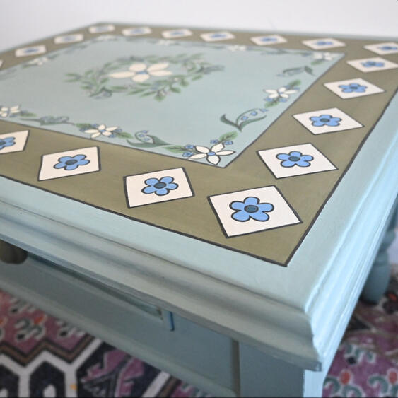 Hand painted wooden table Close-up of a hand-painted floral tabletop featuring blue forget-me-not flowers, white blossoms, green foliage, and a decorative geometric border on a vintage duck egg blue painted wooden table.