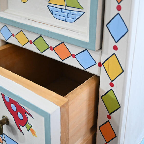 Close-up of an open drawer in a hand-painted children’s wooden storage unit, showing the natural wood interior and colourful geometric diamond border. The drawer front features a red rocket design with a brass knob.
