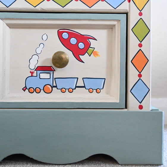 Close-up of a hand-painted children’s wooden drawer featuring a rocket and train, surrounded by a geometric diamond border in orange, blue, yellow and green.