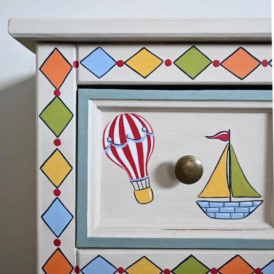 Childs bedside table close up Close-up of a hand-painted children’s wooden drawer featuring a red-and-white hot-air balloon and colourful sailboat, surrounded by a geometric diamond border in orange, blue, yellow and green.