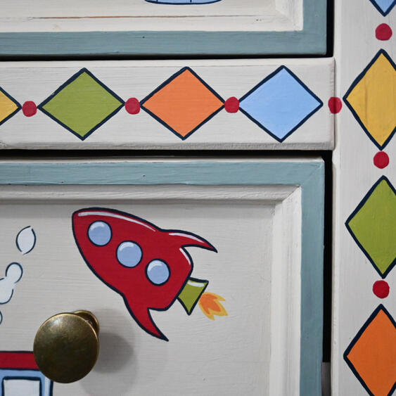 Painted rocket on bedside table Close-up of a hand-painted children’s wooden drawer featuring a rocket surrounded by a geometric diamond border in orange, blue, yellow and green.