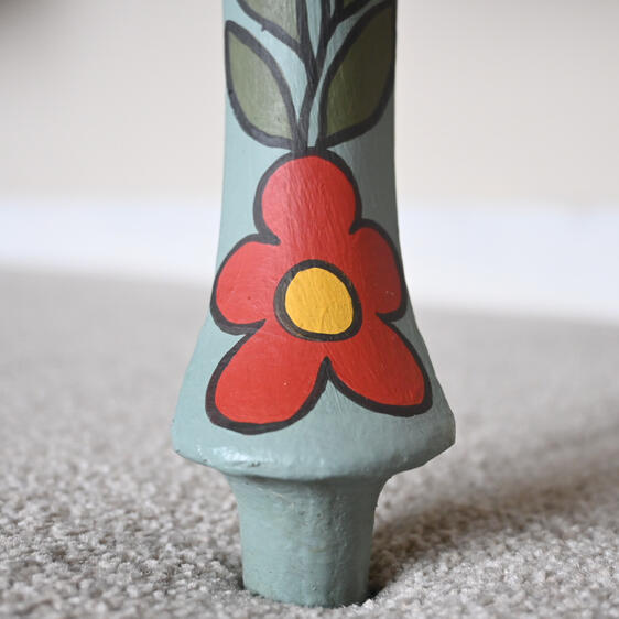Close-up of a painted wooden furniture leg featuring a red flower with a yellow center and green leaves outlined in black, set against a light blue background, resting on beige carpet.