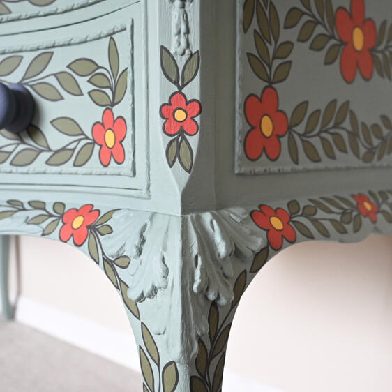 Painted wooden dresser with two drawers, finished in muted teal and decorated with hand-painted red flowers with yellow centers and green leaves. The dresser has ornate carved details, dark round drawer knobs, and curved legs, placed against a light-colore