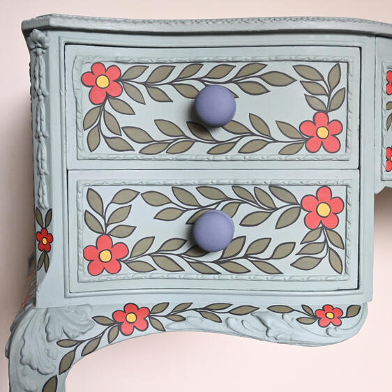 A light blue painted wooden dresser with two drawers, decorated with hand-painted floral patterns. Each drawer has leafy vines and red flowers with yellow centers, while the drawer knobs are round and painted purple. The floral design also extends to the s