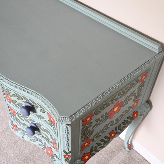 Painted wooden dresser with two drawers, finished in muted teal and decorated with hand-painted red flowers with yellow centers and green leaves. The dresser has ornate carved details, dark round drawer knobs, and curved legs, placed against a light-colore