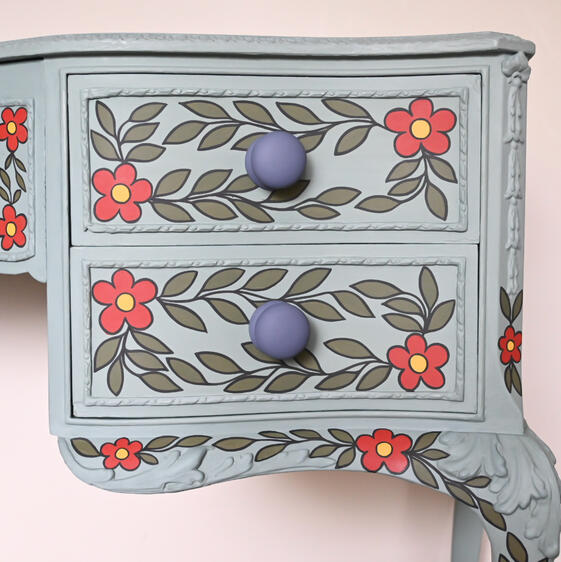 A light blue painted wooden dresser with two drawers, decorated with hand-painted floral patterns. Each drawer has leafy vines and red flowers with yellow centers, while the drawer knobs are round and painted purple. The floral design also extends to the s