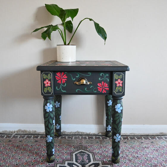 A small dark-painted wooden side table with colorful hand-painted floral designs in red, blue, and green. The table has one drawer with a brass handle