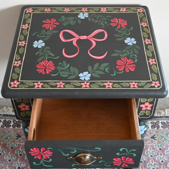 Painted table with open draw A small dark-painted wooden side table with colorful hand-painted floral designs in red, blue, and green. The table has one drawer with a brass handle