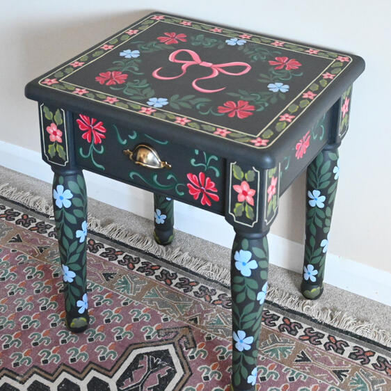 A small dark-painted wooden side table with colorful hand-painted floral designs in red, blue, and green. The table has one drawer with a brass handle