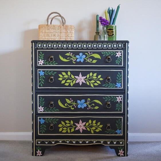 Hand-painted dark wood chest of drawers featuring colorful floral designs, with five drawers and decorative ring handles, styled with a woven basket, candles, and flowers on top against a neutral wall.