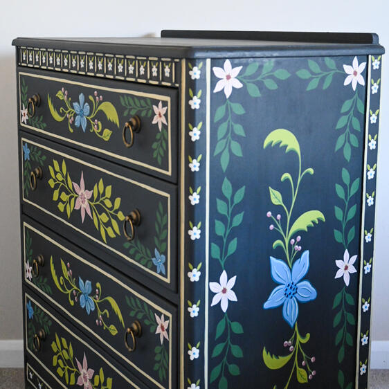 Vintage hand painted cabinet Close-up of a dark hand-painted wooden chest of drawers with colorful floral patterns on the drawers and side panel, featuring decorative ring handles and intricate painted borders.