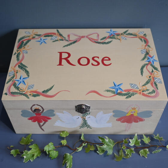 personalised christmas box hand painted personalised Christmas box. Cream wooden keepsake box in cream with a name painted in red. there is a pink ribbon with Christmas foliage surrounding the name and christmas illustrations on the front and side.