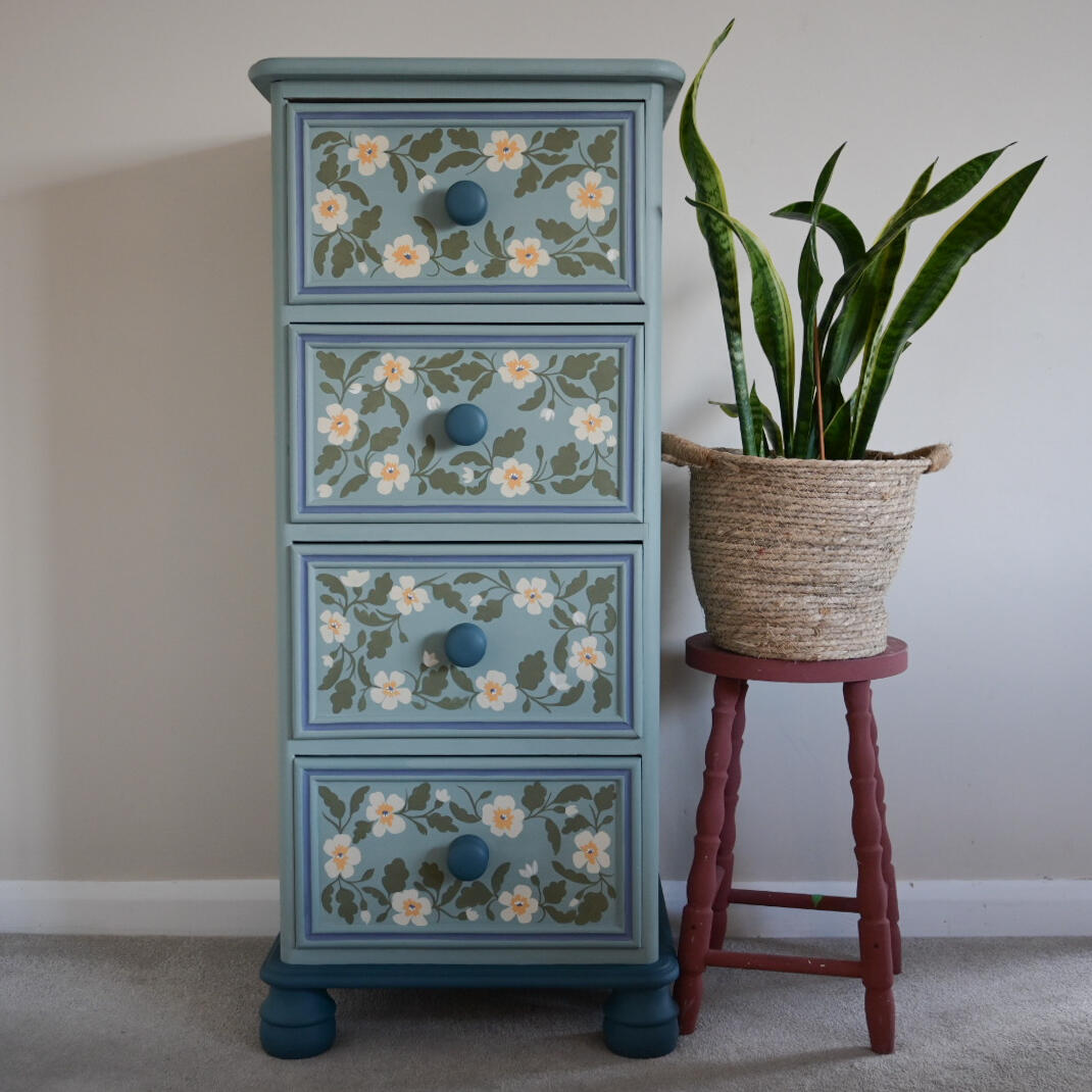Vintage hand painted floral tallboy A tall, narrow chest of drawers painted in a light blue color with hand-painted floral designs on each drawer. The drawers have round blue knobs. A potted snake plant sits to the right of the chest on a small, red wooden stool.