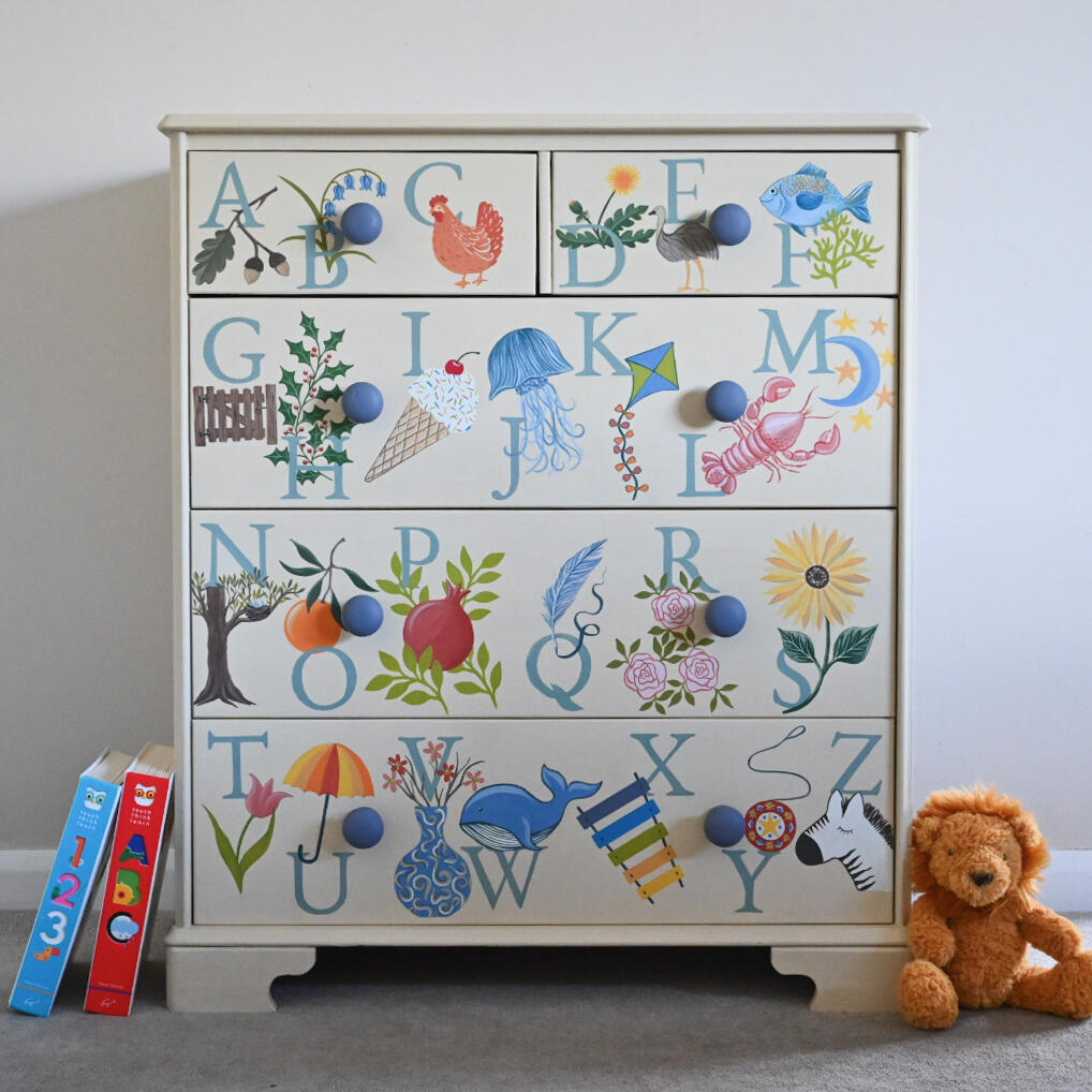 Illustrated alphabet chest of draws A tall, narrow chest of drawers painted in a light blue color with hand-painted floral designs on each drawer. The drawers have round blue knobs. A potted snake plant sits to the right of the chest on a small, red wooden stool.