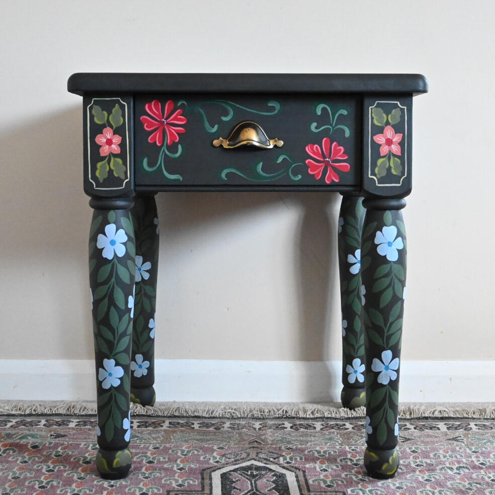 Graphite folky floral side table with draw A small dark-painted wooden side table with colorful hand-painted floral designs in red, blue, and green. The table has one drawer with a brass handle